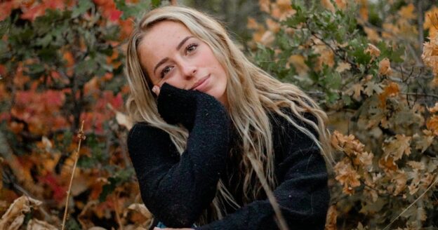 Woman with blonde hair sitting outdoors with fall foliage behind her